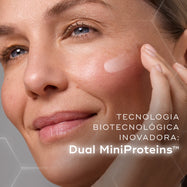 Primeiro plano de uma modella sorridente a aplicar uma porção de creme branco na bochecha contra um fundo com gráficos hexagonais subtis. O texto em português destaca a inovadora tecnologia biotecnológica: Dual MiniProteins™.