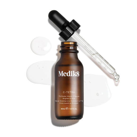 Uma imagem de produto vista de cima (“packshot”) de um frasco de 30 mL do sérum Medik8 C-Tetra  Vitamin C Serum. O frasco de vidro âmbar está sem tampa e colocado ao centro, com a tampa preta do conta-gotas apoiada na diagonal atrás do frasco. Um sérum líquido transparente está artisticamente espalhado e acumulado sobre o fundo branco limpo à volta do frasco, com algumas gotas visíveis na pipeta de vidro. A iluminação é intensa, criando sombras suaves à esquerda e uma estética clínica e elegan-12