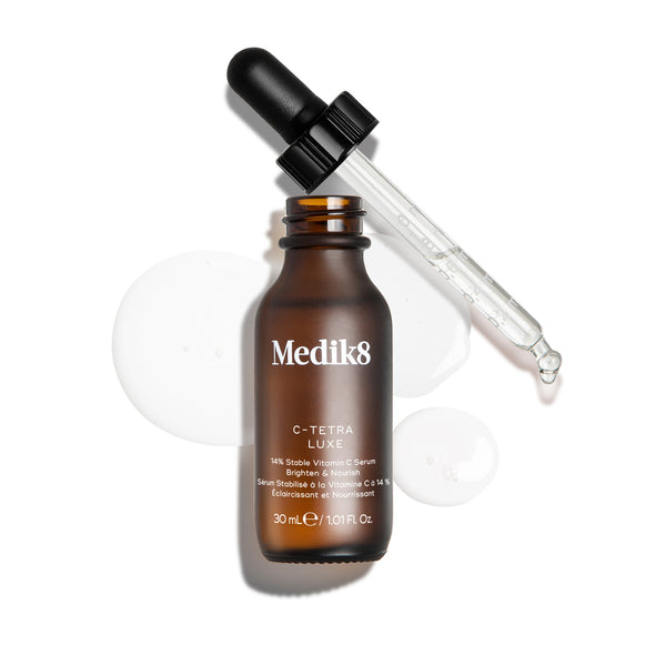 Frasco do sérum Medik8 C-Tetra Luxe apresentado aberto, com a pipeta de vidro apoiada na borda, sobre um fundo branco com gotas de sérum transparentes e translúcidas.-9