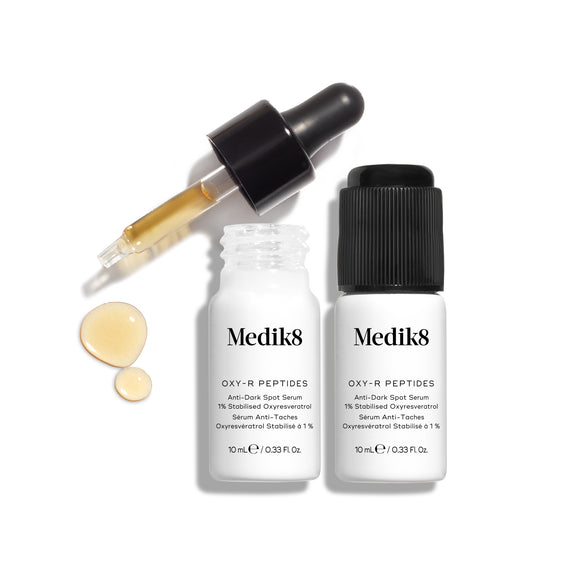 Duas pequenas embalagens brancas do sérum Medik8 Oxy-R Peptides apresentadas. Um conta-gotas preto está colocado ao lado, e duas gotas de sérum amarelo são visíveis sobre a superfície branca.-20